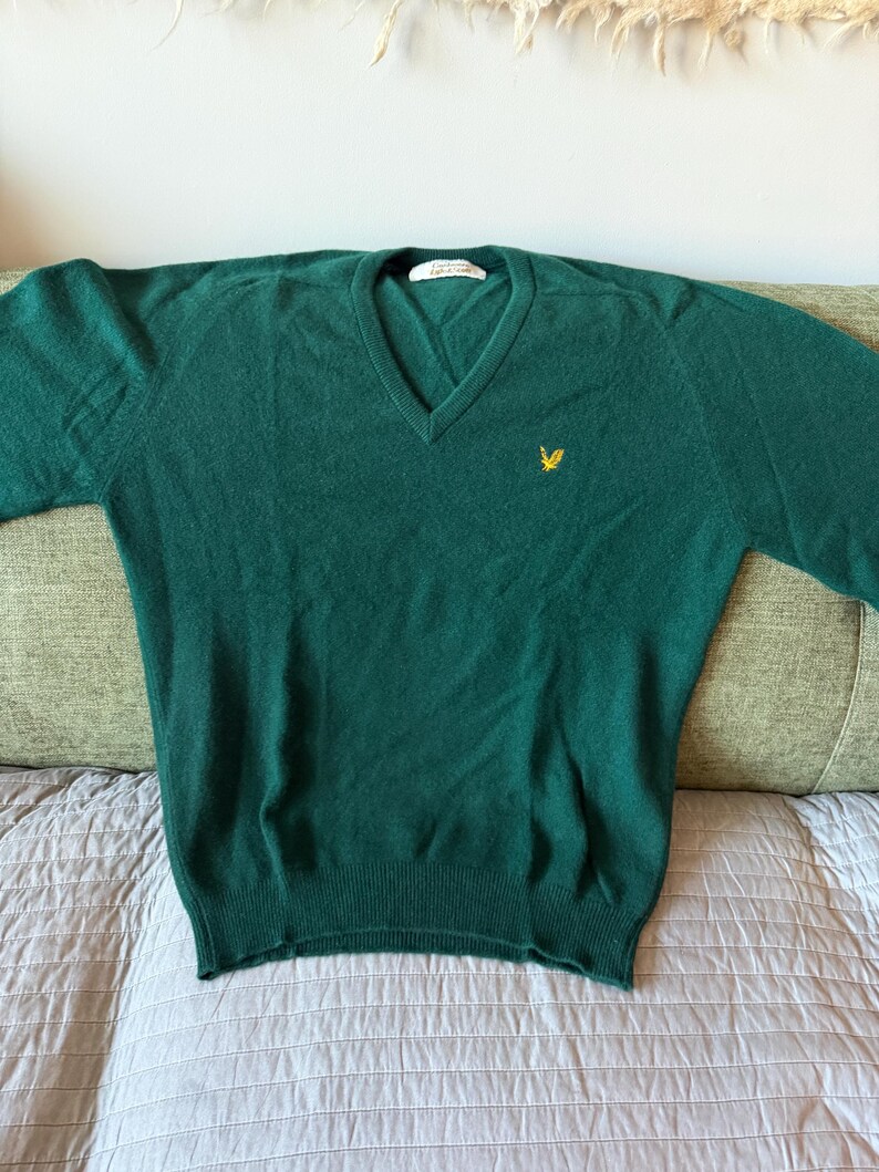 Puede incluir: Un su&eacute;ter de color verde oscuro con cuello en V, mangas largas y dobladillo y pu&ntilde;os acanalados. La prenda presenta un peque&ntilde;o logotipo de &aacute;guila amarilla bordado en el pecho izquierdo y una etiqueta interior blanca con el texto Lyle & Scott.
