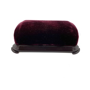 May include: A burgundy velvet jewelry box with a dark wooden base. The box has a rectangular shape with rounded edges and a plush, textured surface. The base has decorative detailing.