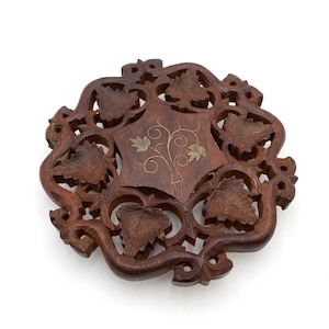 May include: A dark brown, intricately carved wooden coaster with a floral design. The coaster features a central square with a tree and leaf design, accented with gold-colored metal. The outer edge is shaped with carved leaves.