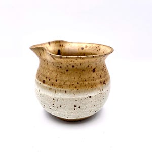 May include: A handmade ceramic creamer with a tan and white speckled glaze. The creamer has a rounded body, a pouring spout, and a wide opening. The top portion is tan, while the bottom is white with brown speckles.