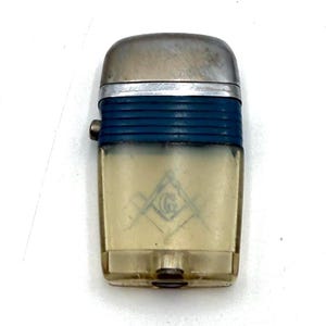 May include: Vintage lighter with a silver top and a blue band. The lower portion is translucent with a Masonic symbol. The lighter has a small button on the side and a metal base. A collectible item.