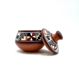 May include: A terracotta lidded bowl featuring a hand-painted band of geometric patterns in black, white, orange, and purple. The bowl has a rounded base and a matching lid with a central knob handle, both decorated with repeating angular motifs.