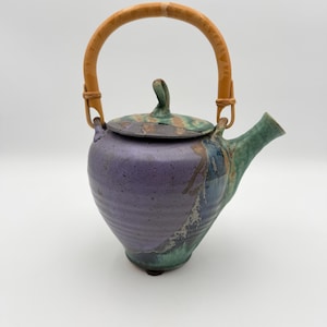 May include: A ceramic teapot featuring a rounded body with a purple and green glaze finish. The teapot has a curved spout, a matching lid with a small handle, and a tall, arched handle made of light brown material attached with woven accents.