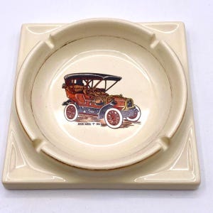 May include: A square ceramic ashtray with a raised rim and a depiction of a red 1903 Buick Model 'C' automobile in the center. The ashtray is cream colored with gold accents.