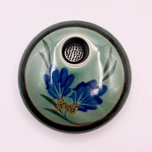 May include: A small, round ceramic vase with a green glaze and a blue floral design. The vase has a small opening at the top for flowers.