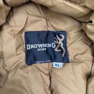 May include: A tan quilted down jacket featuring a black rectangular interior label with the white text Browning Down, a gold buckmark logo, and the text Made in USA. A small white tag below the label indicates the size as XL.