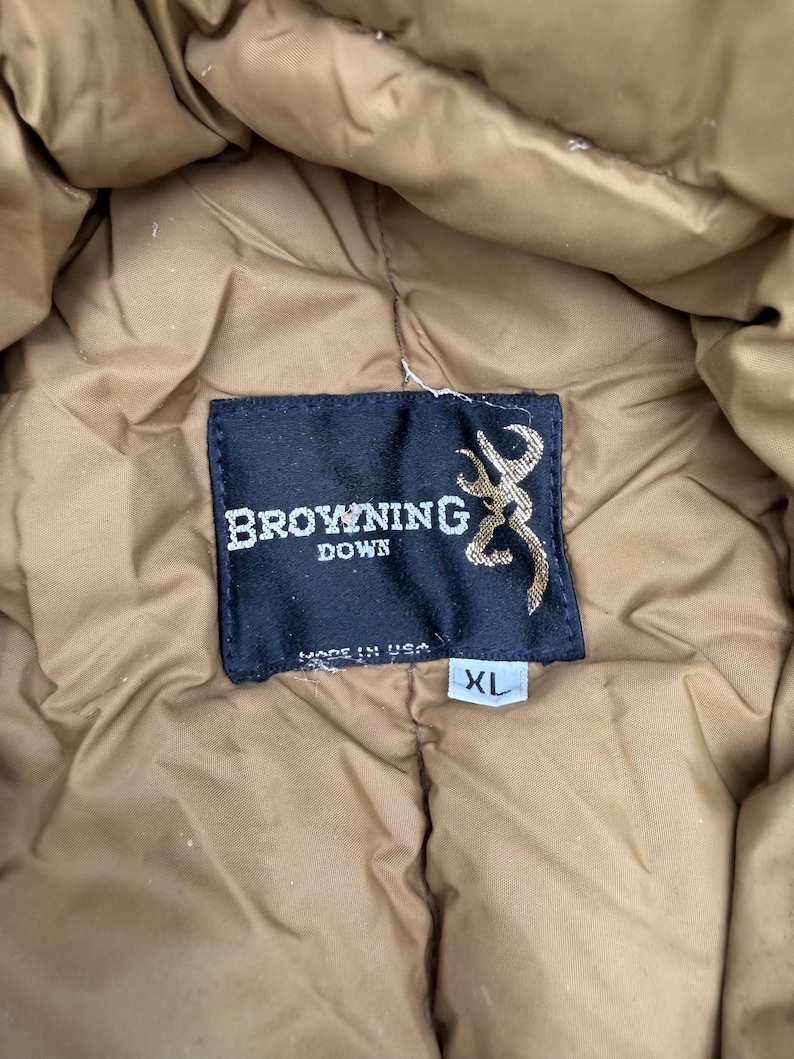 May include: A tan quilted down jacket featuring a black rectangular interior label with the white text Browning Down, a gold buckmark logo, and the text Made in USA. A small white tag below the label indicates the size as XL.