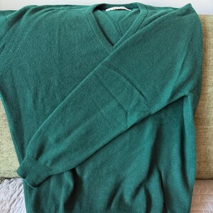 Puede incluir: Un su&eacute;ter de manga larga en color verde oscuro s&oacute;lido con cuello en V, pu&ntilde;os acanalados en las mu&ntilde;ecas y un dobladillo acanalado en la parte inferior. La prenda est&aacute; confeccionada en un tejido de punto suave y tiene una etiqueta blanca.