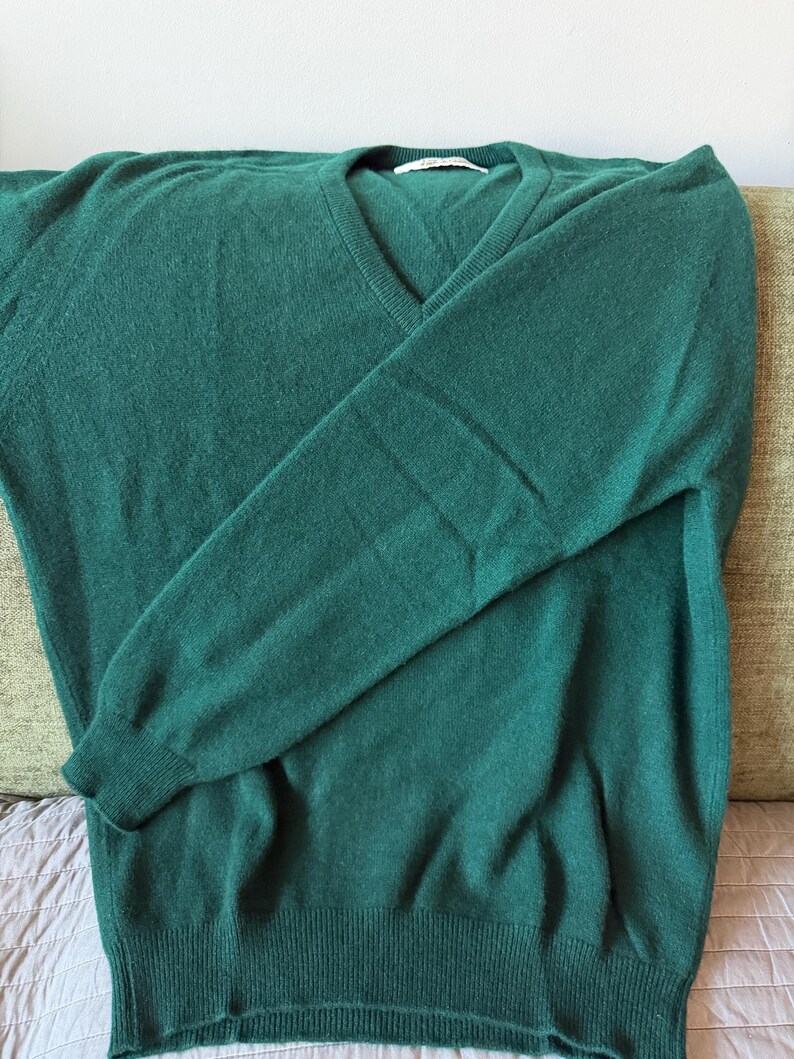 Puede incluir: Un su&eacute;ter de manga larga en color verde oscuro s&oacute;lido con cuello en V, pu&ntilde;os acanalados en las mu&ntilde;ecas y un dobladillo acanalado en la parte inferior. La prenda est&aacute; confeccionada en un tejido de punto suave y tiene una etiqueta blanca.