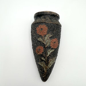 May include: A black ceramic wall vase with a textured surface and a floral design. The vase has a dark brown rim and features three orange flowers with green and gray leaves. The vase is shaped like an arrow.