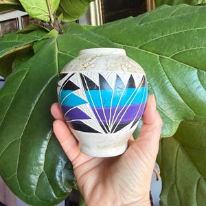 May include: A small, hand-painted ceramic vase with a cream-colored base and a colorful geometric design. The vase features black, turquoise, and purple shapes, with white lines. The vase is held in a hand, with a large green plant in the background.