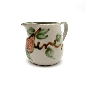 May include: Cream-colored ceramic pitcher with a handle and spout. The pitcher is decorated with a brown vine, green leaves, and a reddish-orange pear. The word "June" is written on the pitcher in brown script.