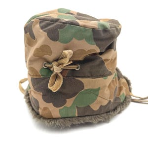 May include: A camouflage print hat featuring a quilted exterior with brown, tan, and green patterns. The hat includes a tan fabric drawstring tied in a bow through metal eyelets and a thick, dark gray faux fur lining along the bottom edge.