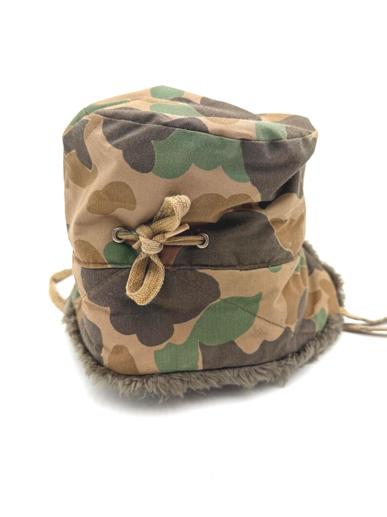 May include: A camouflage print hat featuring a quilted exterior with brown, tan, and green patterns. The hat includes a tan fabric drawstring tied in a bow through metal eyelets and a thick, dark gray faux fur lining along the bottom edge.