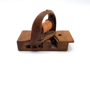May include: A vintage wooden thread holder with a spool of orange thread. The holder has a rectangular base and a curved handle. The wood has a dark, aged appearance, suggesting it's an antique or a well-used item. The design is simple and functional.