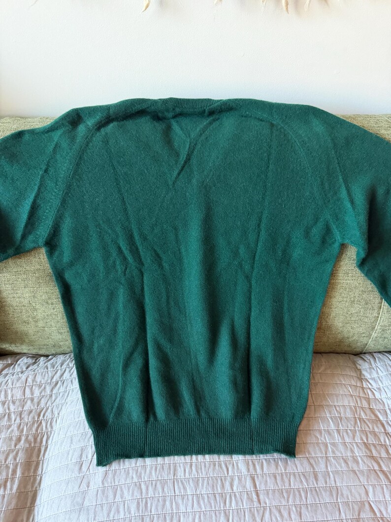 Puede incluir: Un su&eacute;ter de manga larga en color verde oscuro s&oacute;lido con cuello en V, pu&ntilde;os acanalados y dobladillo acanalado. La prenda est&aacute; confeccionada con un tejido de punto suave y texturizado, presentando un dise&ntilde;o cl&aacute;sico con mangas ragl&aacute;n.