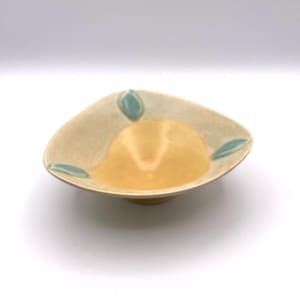 May include: A handmade ceramic bowl with a unique, organic shape. The bowl features a light beige exterior with a yellow interior. Teal leaf-shaped accents are placed around the rim. The bowl is approximately 8 inches in diameter.