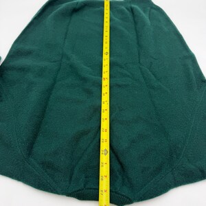 Puede incluir: Una falda de punto verde oscuro con cintura acanalada y detalles de costuras verticales. Una cinta m&eacute;trica amarilla est&aacute; colocada verticalmente sobre el centro, indicando una longitud total de unos 73,5 cm desde la cintura hasta el dobladillo.