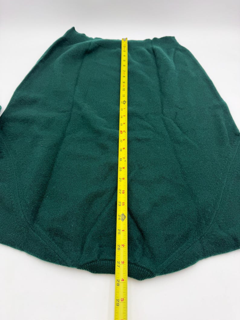 Puede incluir: Una falda de punto verde oscuro con cintura acanalada y detalles de costuras verticales. Una cinta m&eacute;trica amarilla est&aacute; colocada verticalmente sobre el centro, indicando una longitud total de unos 73,5 cm desde la cintura hasta el dobladillo.