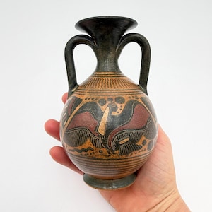 May include: A small, antique-style ceramic vase with a dark brown rim and handles. The vase features a tan body with black and maroon geometric designs and horizontal stripes. The vase is held in a hand.