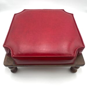 May include: A red, square ottoman with a padded top and a wooden base. The ottoman has a dark wood base with four turned legs. The red top has a decorative edge.