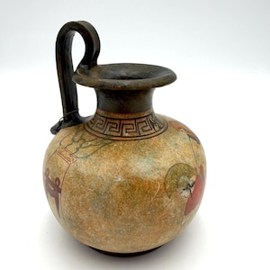 May include: A small, antique-style ceramic vase with a dark handle and rim. The vase has a rounded body with painted figures in red, green, and brown. The neck features a decorative geometric pattern.