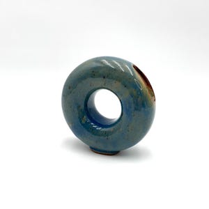 May include: A ceramic vase with a circular design and a hole in the center. The vase is glazed in shades of blue and green, with a reddish-brown interior. The vase has a smooth, glossy finish.