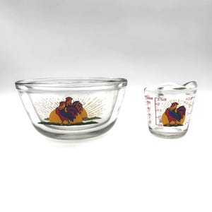 May include: Clear glass mixing bowl and measuring cup set. Both pieces feature a colorful rooster design with a yellow sunburst background. The measuring cup has volume markings in cups and milliliters.