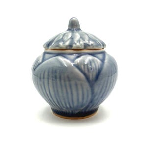 May include: A small, blue ceramic jar with a lid. The jar has a rounded shape with a textured, petal-like design. The lid has a small knob on top. The jar is on a white background.