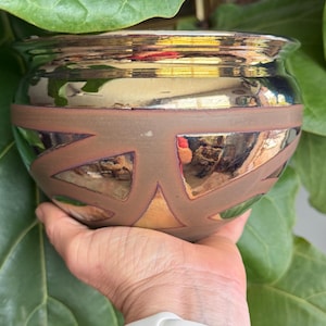 May include: A round ceramic bowl featuring a metallic, reflective finish on the upper and lower sections, contrasted by a matte, earthy brown band around the center. The bowl displays a geometric pattern of triangular shapes across its surface.