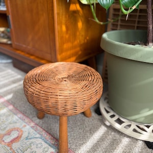 May include: A small, round stool featuring a woven wicker seat in a natural light brown tone. The stool is supported by three sturdy, smooth wooden legs that extend outward from the base, providing a stable foundation for the circular top.