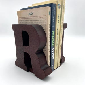 May include: A dark brown wooden bookend shaped like the letter "R" supports several books. The books have various colored spines and titles, including "Music Lust" and "The Cloudspotter's Guide".
