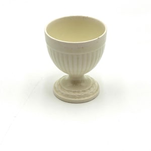 May include: Off-white ceramic egg cup with a fluted body and a round, pedestal base. The egg cup has a smooth, slightly glossy finish and a simple, classic design. The interior is a pale yellow color.