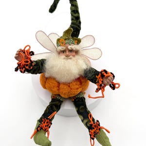 May include: A whimsical fairy figurine with a long, pointed green velvet hat, a fluffy white beard, and a round orange pumpkin-shaped body. The figure features translucent wings, green patterned limbs, and bright orange decorative wire accents.