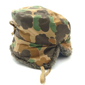 May include: A camouflage pattern winter hat featuring a rounded crown and ear flaps lined with dark gray faux fur. The exterior fabric displays a brown, tan, and green cloud-like camouflage print. A tan fabric tie hangs from the bottom edge.