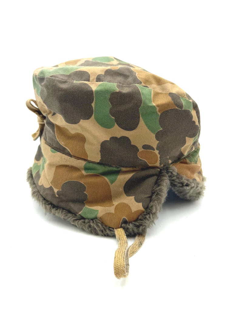 May include: A camouflage pattern winter hat featuring a rounded crown and ear flaps lined with dark gray faux fur. The exterior fabric displays a brown, tan, and green cloud-like camouflage print. A tan fabric tie hangs from the bottom edge.