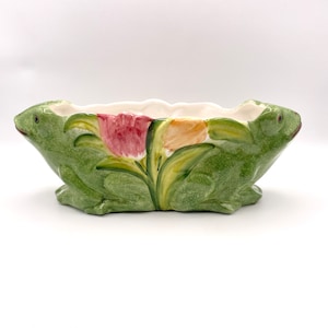 May include: A green ceramic planter shaped like two frogs back to back. The planter has a floral design with pink, yellow, and green tulips.