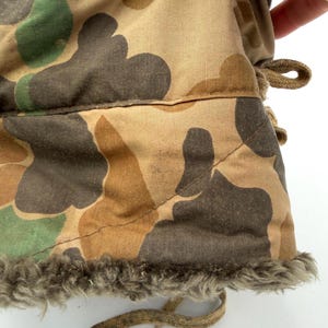 May include: A camouflage patterned garment featuring a tan, brown, and green organic shape print. The fabric has a quilted texture with visible stitching and a thick, textured gray faux fur lining along the bottom edge, accompanied by tan fabric ties.