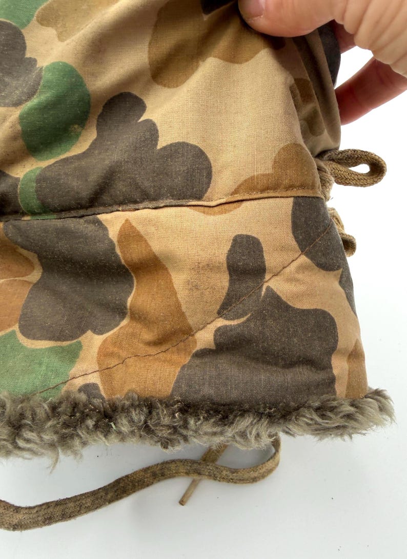 May include: A camouflage patterned garment featuring a tan, brown, and green organic shape print. The fabric has a quilted texture with visible stitching and a thick, textured gray faux fur lining along the bottom edge, accompanied by tan fabric ties.
