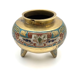 May include: A small, round, brass-colored vessel with three small feet. The body is decorated with a band of blue, red, and white enamel floral designs. The top rim is brass-colored, and the interior is dark.