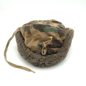 May include: A camouflage print trapper hat featuring a brown, green, and tan pattern. The hat is lined with thick, textured brown faux fur and includes a tan drawstring cord with metal eyelets at the center for an adjustable fit.