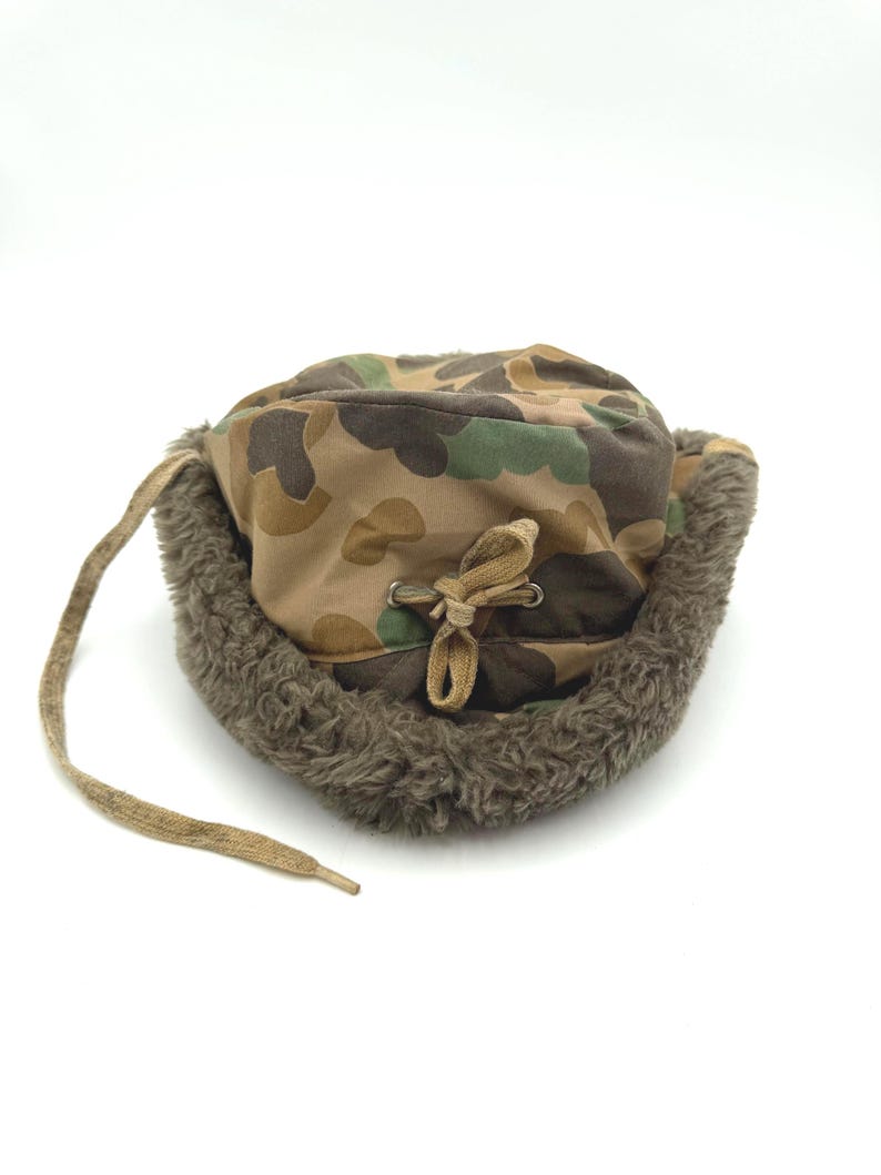 May include: A camouflage print trapper hat featuring a brown, green, and tan pattern. The hat is lined with thick, textured brown faux fur and includes a tan drawstring cord with metal eyelets at the center for an adjustable fit.