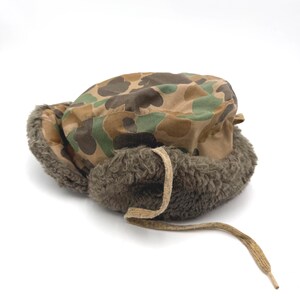 May include: A camouflage print trapper hat featuring a brown, green, and tan pattern on the crown. The hat includes thick, dark olive-brown faux fur lining along the ear flaps and a single tan fabric tie hanging from the front of the brim.