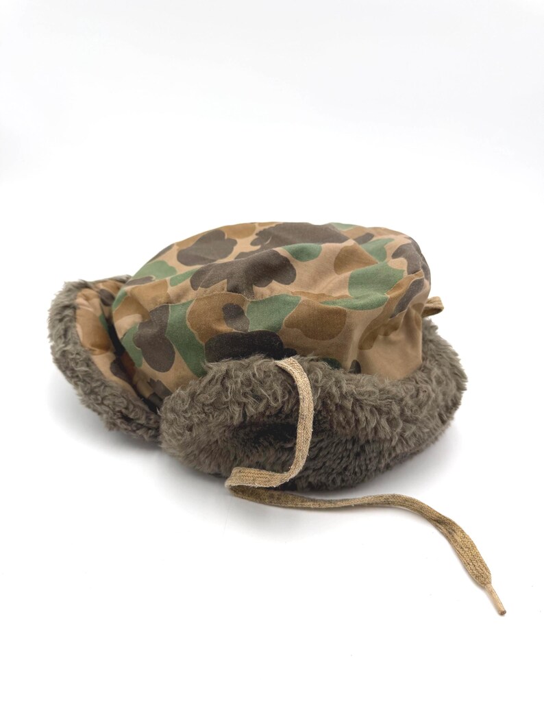 May include: A camouflage print trapper hat featuring a brown, green, and tan pattern on the crown. The hat includes thick, dark olive-brown faux fur lining along the ear flaps and a single tan fabric tie hanging from the front of the brim.