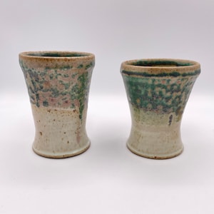 May include: Two ceramic cups with a white glaze and green and pink speckled designs. The cups have a flared rim and a rounded base.