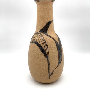 May include: A tan ceramic vase with a narrow neck and rounded body. Dark blue abstract lines and shapes decorate the vase. The rim of the vase is dark blue.
