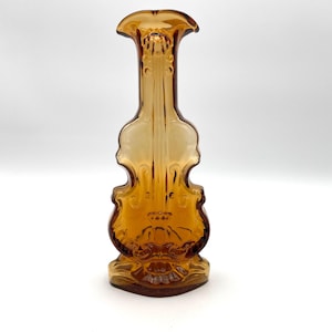 May include: Amber-colored glass vase in the shape of a violin. The vase has a flared top and a detailed design on the front, including the violin's curves and decorative elements. The base is also detailed, and the vase is made of transparent glass.