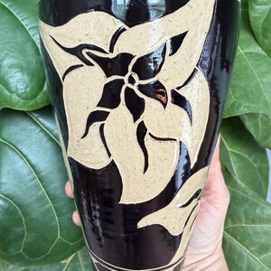 May include: A tall, tapered ceramic vase featuring a glossy dark brown glaze with a contrasting matte beige sgraffito floral design. The vessel has a wide, open top that narrows toward the base, decorated with a geometric band near the bottom edge.