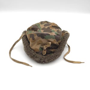 May include: A camouflage print trapper hat featuring a brown, green, and tan pattern. The hat includes a thick, dark brown faux fur lining along the brim and ear flaps, with two tan fabric ties hanging from the sides and a central drawstring.