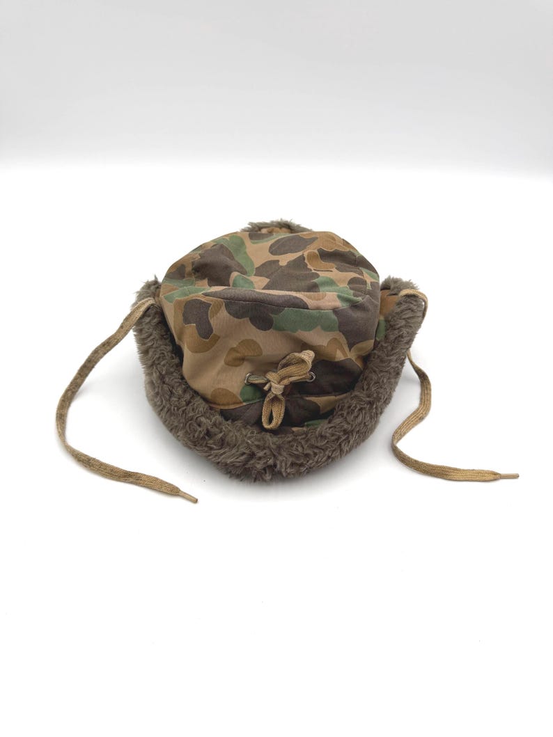 May include: A camouflage print trapper hat featuring a brown, green, and tan pattern. The hat includes a thick, dark brown faux fur lining along the brim and ear flaps, with two tan fabric ties hanging from the sides and a central drawstring.